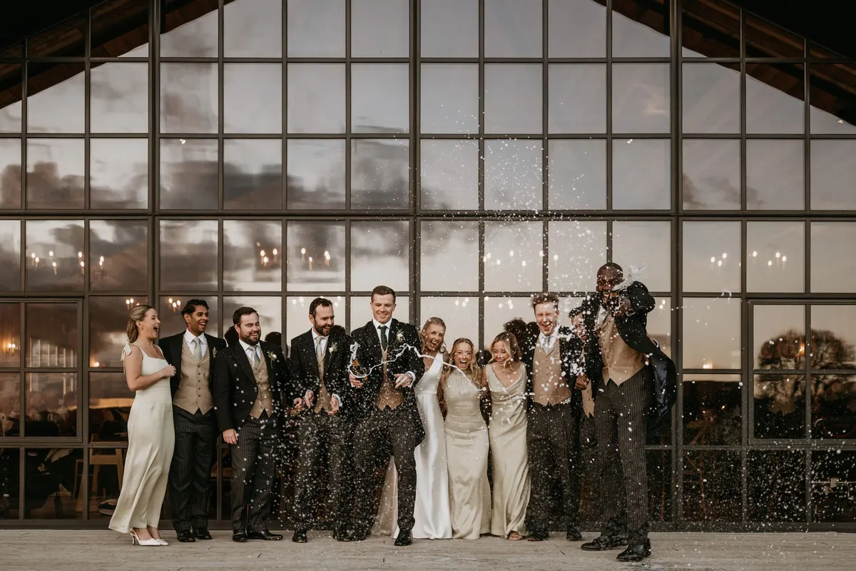 A wedding party laughing outside Botley Hill Barn as a champagne cork goes off, the glass gable of the barn behind them reflecting the evening sky and the candlelit tables inside