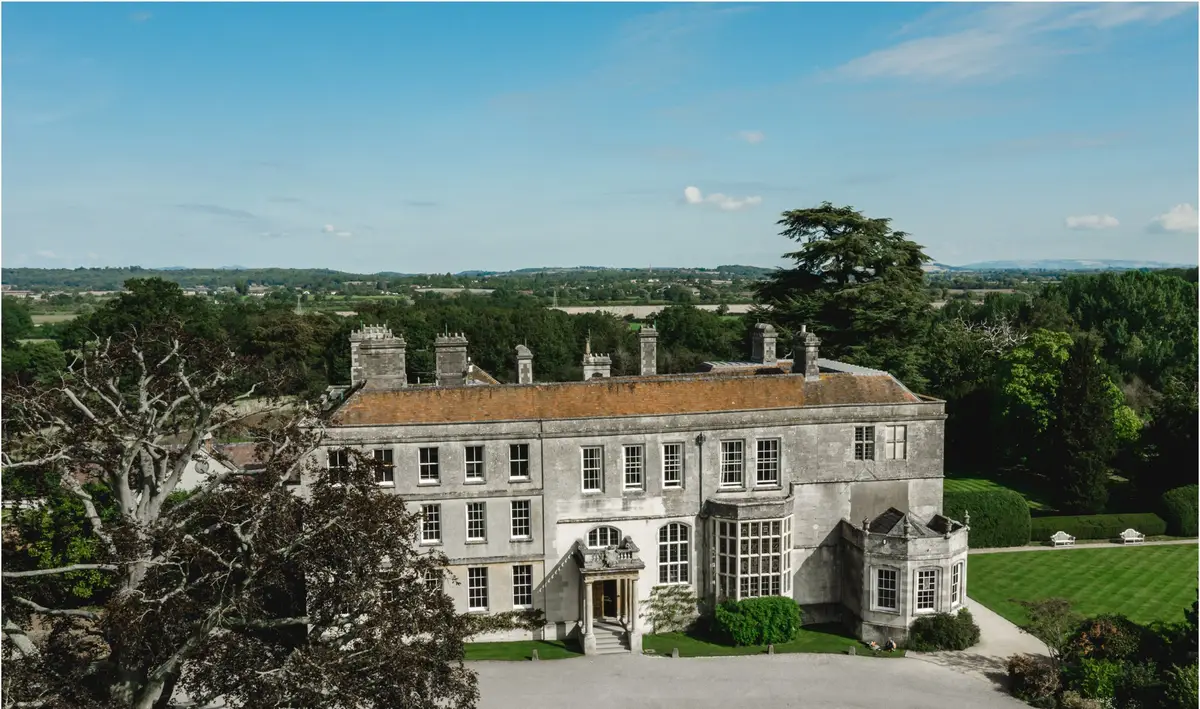 A Cotswold stone manor house seen from above, with a cedar tree to one side and open countryside behind