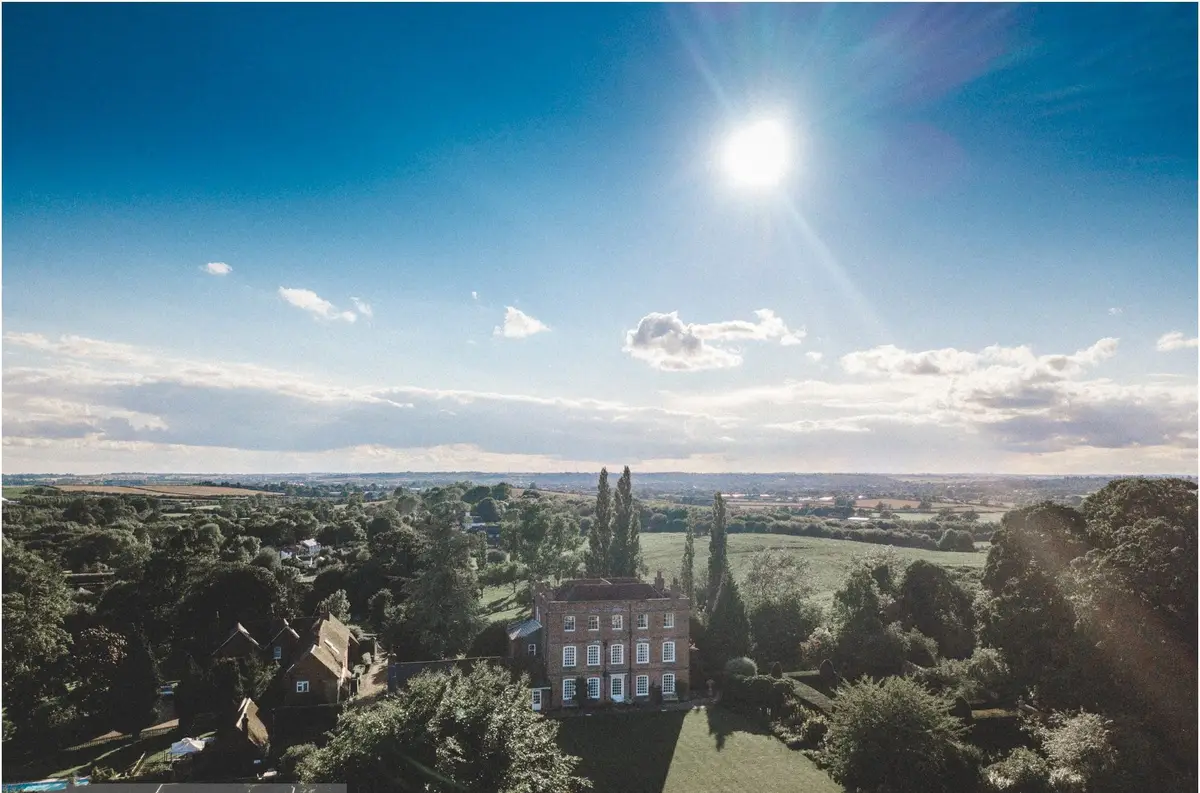 A Georgian country house and its gardens from the air under a bright sky with scattered cloud, fields and hedgerows stretching to the horizon