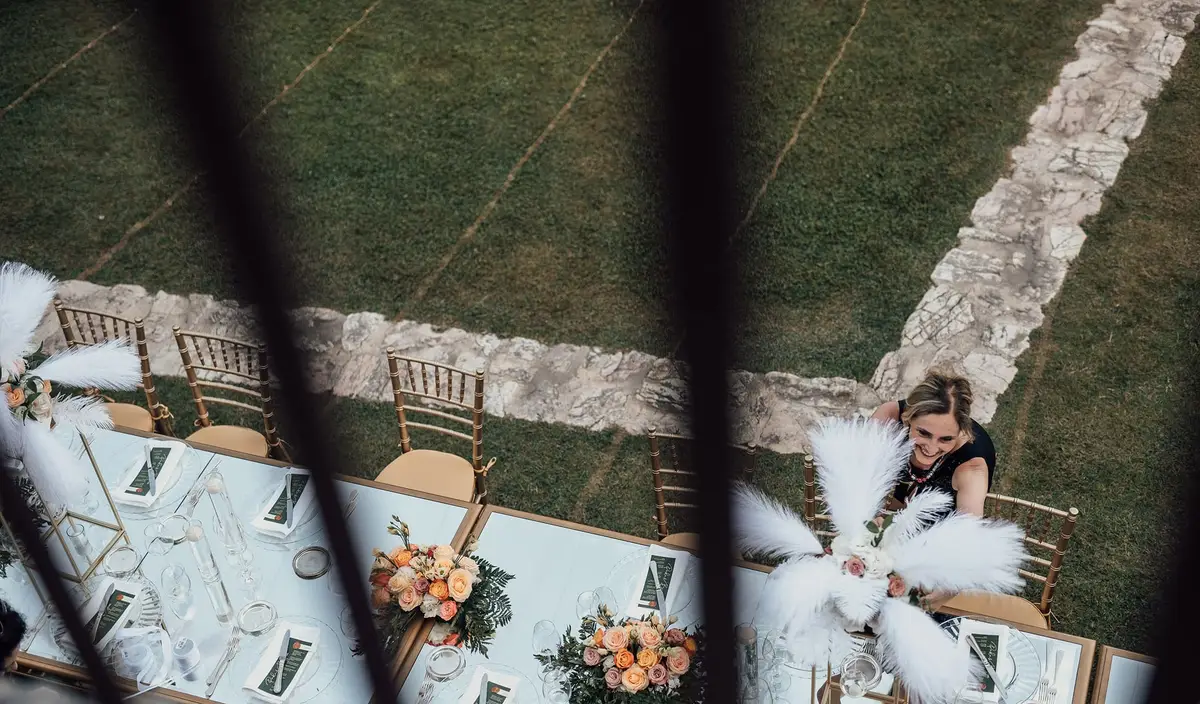 A long outdoor table on a lawn seen from above, laid with candles and flowers, with a bride adjusting her dress at one end