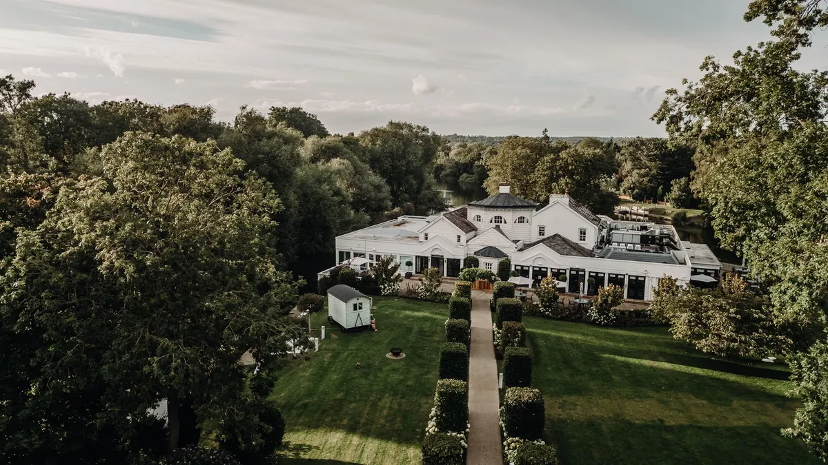 Monkey Island Estate from the air: a white riverside house with a domed pavilion among mature trees on the Thames, a clipped hedge walk leading across the lawn to its terrace