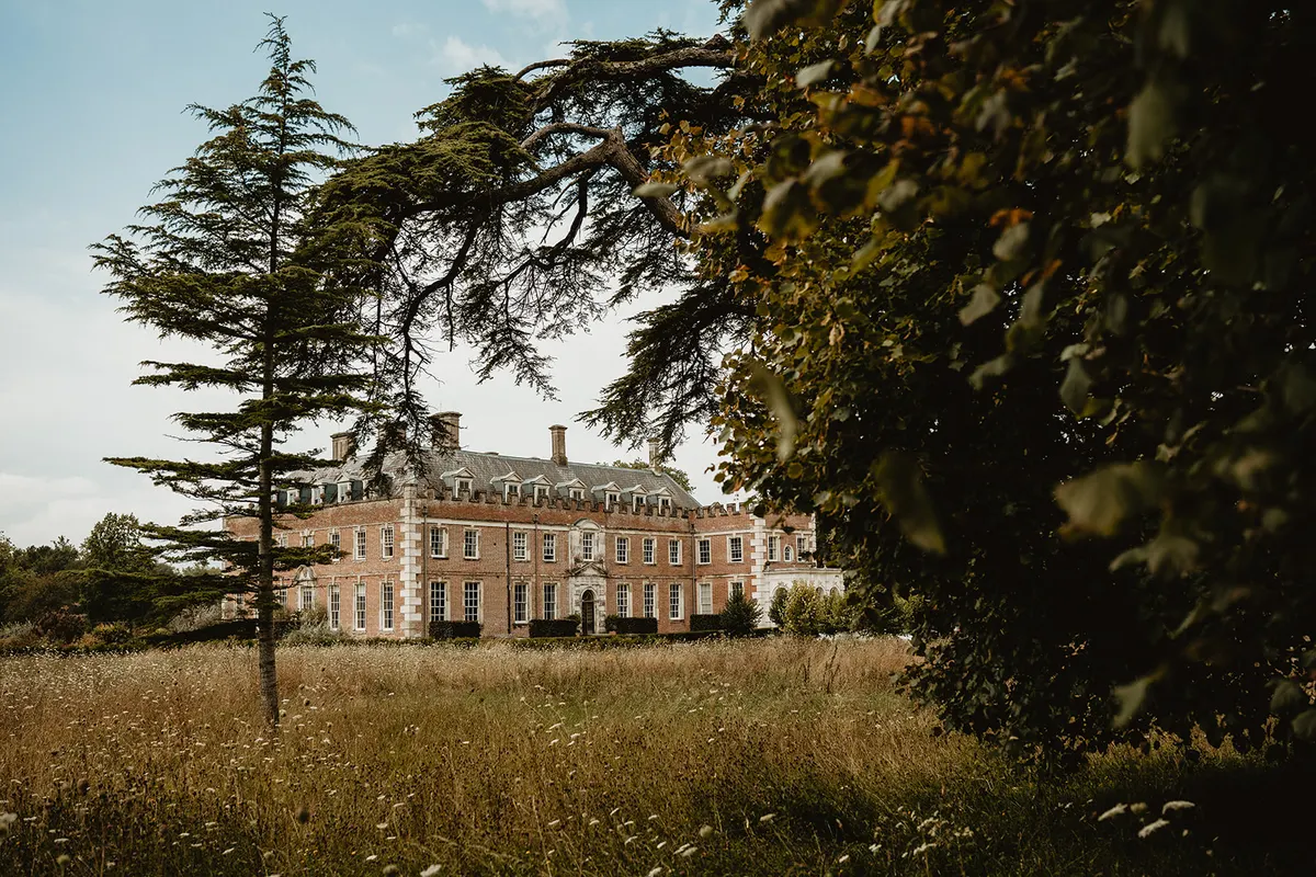 St Giles House seen across a wildflower meadow, a red-brick country house with white stone dressings framed by the boughs of a cedar and a beech in full leaf