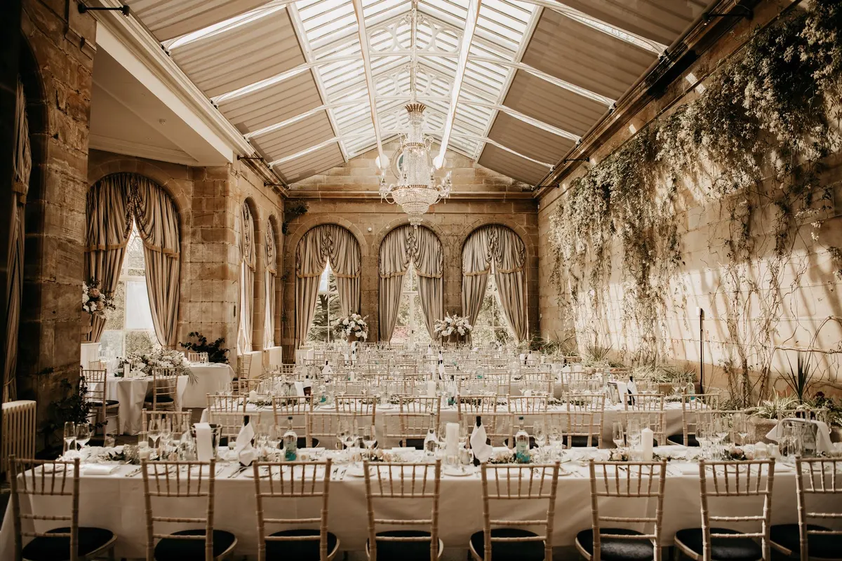The orangery at Weston Park laid for a wedding breakfast: long tables under a glass roof and a crystal chandelier, arched windows with swagged curtains, and a wall of trailing greenery and blossom